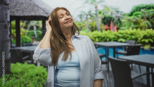 Woman touching hair with hand beside pool and cabana at a tropical resort building overlooking palms and outdoor table; serenity. Free Download Woman touching hair with hand beside pool and cabana at a tropical resort building overlooking palms and outdoor table; serenity. - GFXLoad