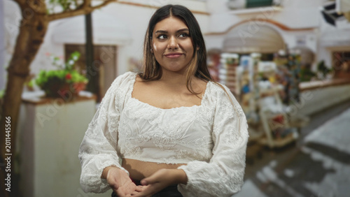 Obraz na płótnie Woman in a white embroidered blouse clasping hands near chest and smiling on a mediterranean street beside a souvenir kiosk and planter; gentle joy