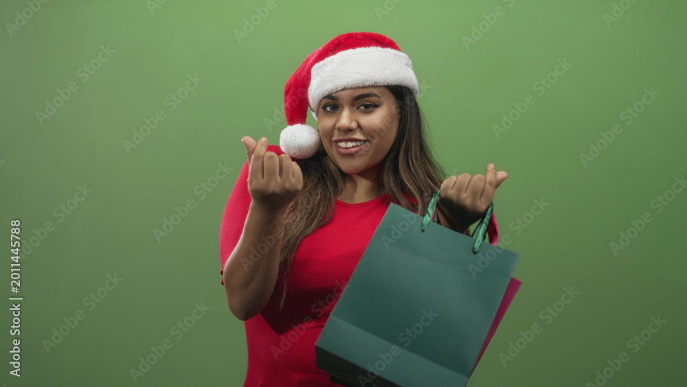 Naklejka premium Woman in santa hat and red dress holds shopping bag and beckons with both hands in studio; holiday joy.
