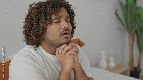 Slika na platnu Young indian man on bed in bedroom, white t shirt, eyes closed with hands clasped in prayer and pleading expression, orange pillow and bedside vases visible; quiet devotion