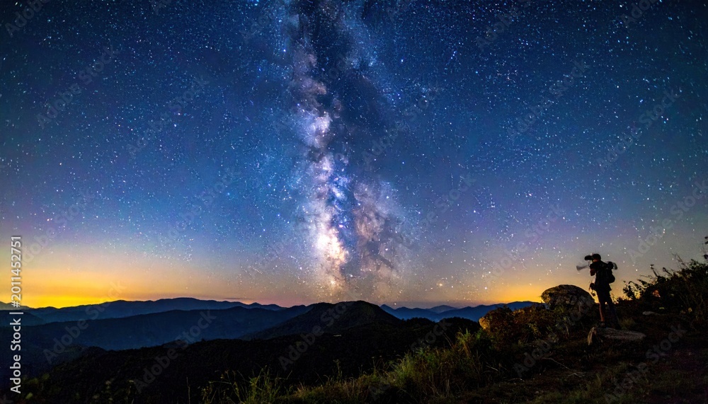Fototapeta premium Starry night sky with Milky Way over silhouetted mountains; person photographing celestial scene