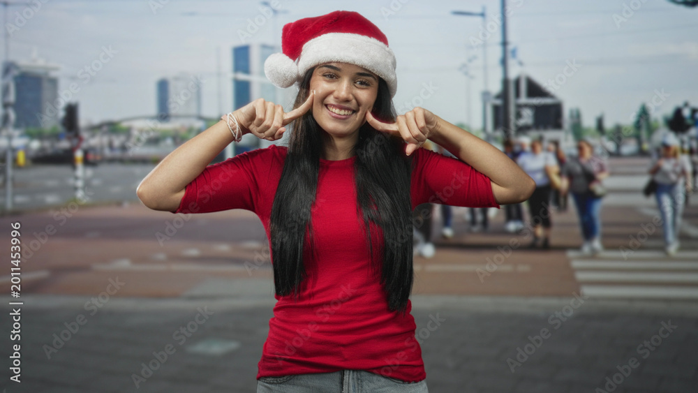 Fototapeta premium Young woman points index fingers to cheeks and smiles in a busy city street crosswalk wearing santa hat and red shirt, long hair visible; holiday joy.