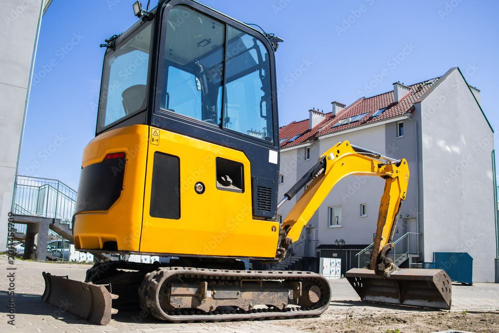 Obraz premium Yellow mini excavator parked on construction site with residential buildings in the background under clear blue sky and bright sunlight