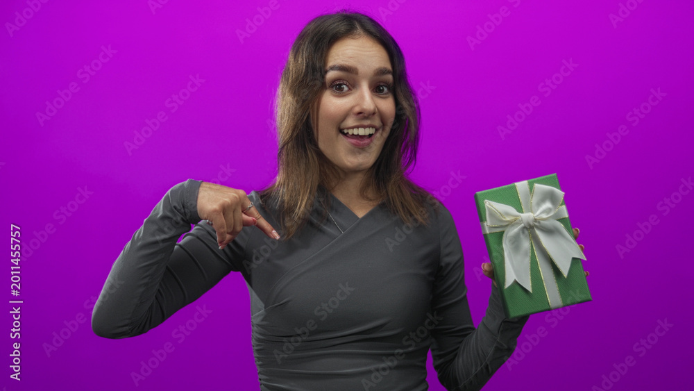 Fototapeta premium Woman pointing finger to green gift box while holding present in magenta studio; celebration surprise joy.