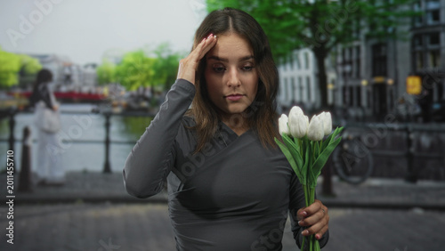 Foto Woman holding white tulips touches her temple while wearing a gray wrap dress on an amsterdam street by a canal; quiet sadness reflection