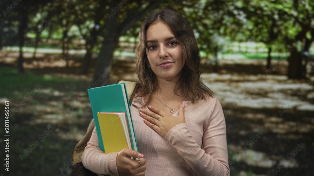 Fototapeta premium Woman holding notebooks and folders, hand on chest and backpack strap visible standing under trees in park; gratitude.