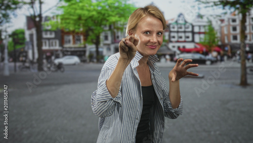 Foto Blond woman in striped shirt shows open palms, spreads fingers and pushes hands toward camera on an urban street framed by canalside houses and trees; playful curiosity