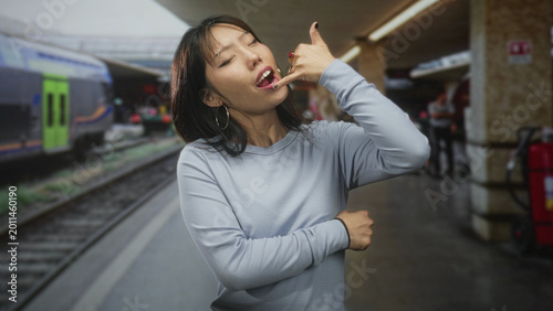 Foto Young chinese woman making phone call hand gesture with crossed arm at train station platform; playful flirtation