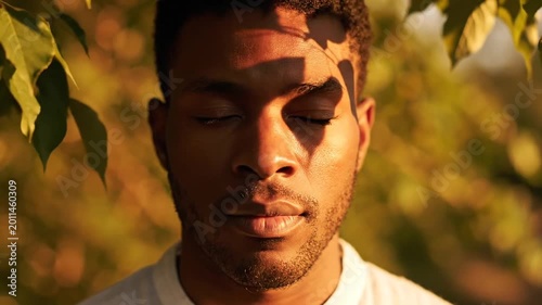 Young African American man with closed eyes bathed in warm sunlight and dappled shadows from green leaves creating a peaceful outdoor portrait