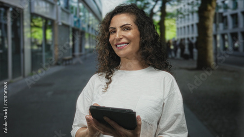 Foto Young hispanic woman holding tablet taps screen with hand while standing on city street; confidence
