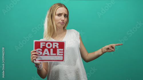 Εκτύπωση καμβά Woman blonde holding a for sale sign and pointing finger to the right in studio with teal backdrop, showing a persuasive selling pose; sale negotiation doubt