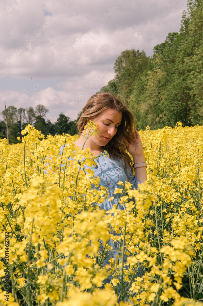 Fototapeta premium A beautiful pregnant woman with long wavy hair, wearing a light blue floral dress, gently holding her belly while standing in a blooming yellow rapeseed field. 