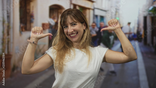 Принти на полотні Woman smiling with thumbs pointing to chest on narrow street with blurred pedestrians and buildings behind her; playful confidence