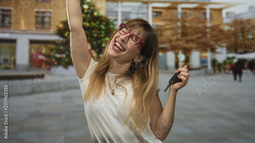 Photos Woman holding car keys and flexing arm in city street next to seasonal decorations; joy celebration