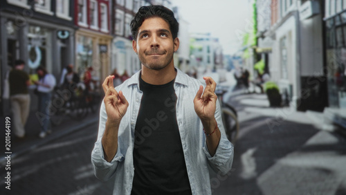 Fotografie Man with hands showing fingers crossed and looking up on a busy street, wearing casual shirt and bracelet; hopeful luck wish