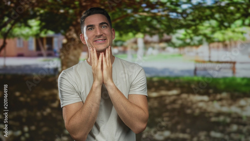 Man with hands on cheeks smiling in forest park near bench and tree in sunlight wearing casual white tshirt; surprise wonder.