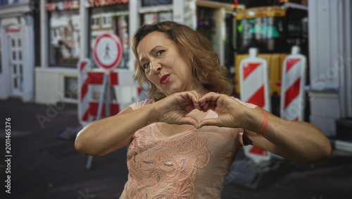 Bilde på lerret Smiling woman shaping heart with her fingers on a sunny city street in front of construction barriers; love