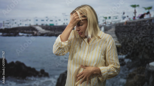 Vászonkép Woman with hand on chest and other hand to forehead near rocky pier and building by the seaside, showing fainting gesture; dizziness concern