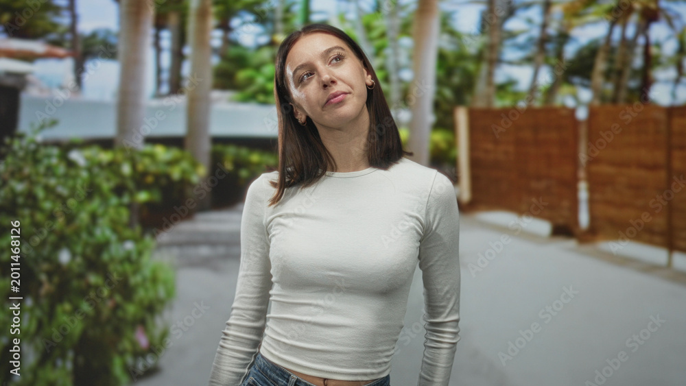 Fototapeta premium Young woman smiling and looking up on a street, wearing a white long sleeve top and jeans with relaxed posture; serene confidence.