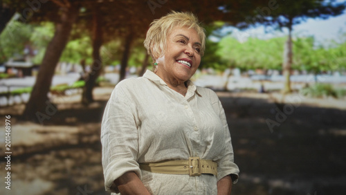 Photos Woman smiling with face and tilted head in park wearing beige linen shirt and belt beneath trees and sunlight; contentment calm