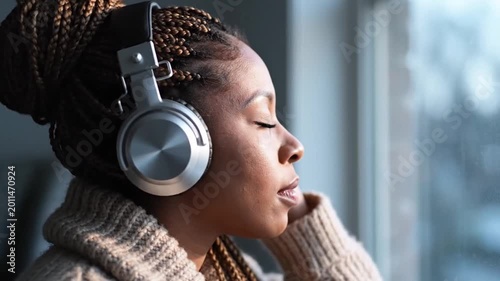 Relaxed African American Woman With Braids Wearing Silver Headphones Listening To Music By A Window During Golden Hour At Home