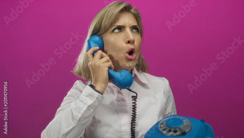Bild auf Leinwand Young blonde woman holding blue phone handset to her ear in vibrant pink studio setting; frustration