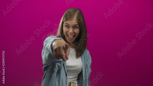 Quadro su tela Young blonde woman pointing finger at camera with finger to lips for silence in pink studio; playfulness