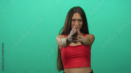 Photos Young hispanic woman pointing fingers like a gun with hands extended in green studio; determination resolve focus strength