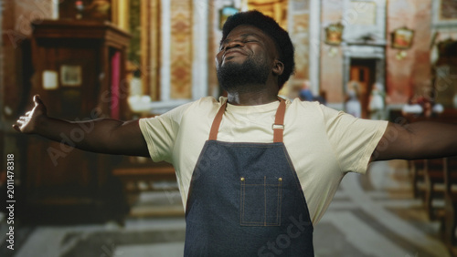 Konstfotografi Man with arms outstretched in church, wearing denim apron and cream t shirt, head tilted upward and eyes closed; faith gratitude joy