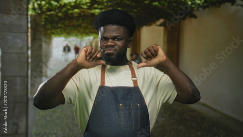 Konstfotografi Man in denim apron points both thumbs at chest while grimacing on a stone street arcade; self judgement disapproval