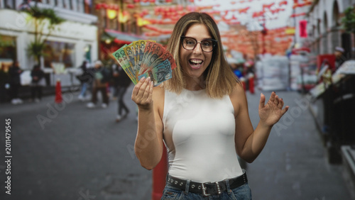 Принти на полотні Young blonde hispanic woman smiling holds fan of australian dollars in right hand on street with lanterns and passersby; joy winning confidence