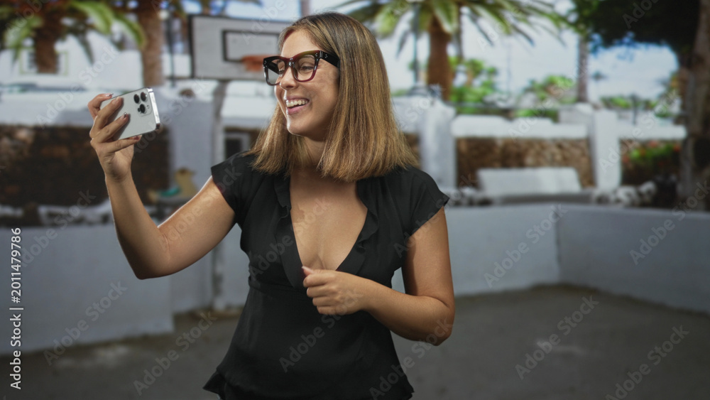 Fototapeta premium Woman holding smartphone with finger to lips gesture while smiling in a street setting with a basketball hoop visible; playful curiosity.