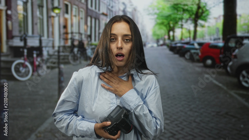 Tableau sur toile Woman holding camera with hand on chest and open mouth, pointing finger while standing on city street with parked cars and bicycles; surprise