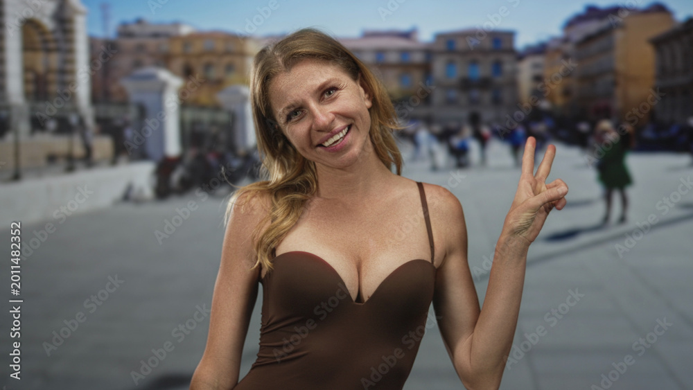 Fototapeta premium Woman smiling and holding up two fingers in a peace sign with her right hand, wearing a brown strap dress with visible cleavage in a busy street plaza; playful confidence.