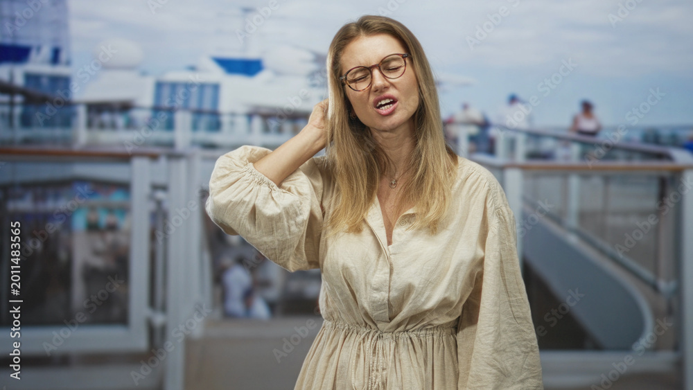 Fototapeta premium Blonde woman holds neck with her hand on a ship deck, eyes closed and wincing, wearing glasses and beige dress near railing; discomfort travel.