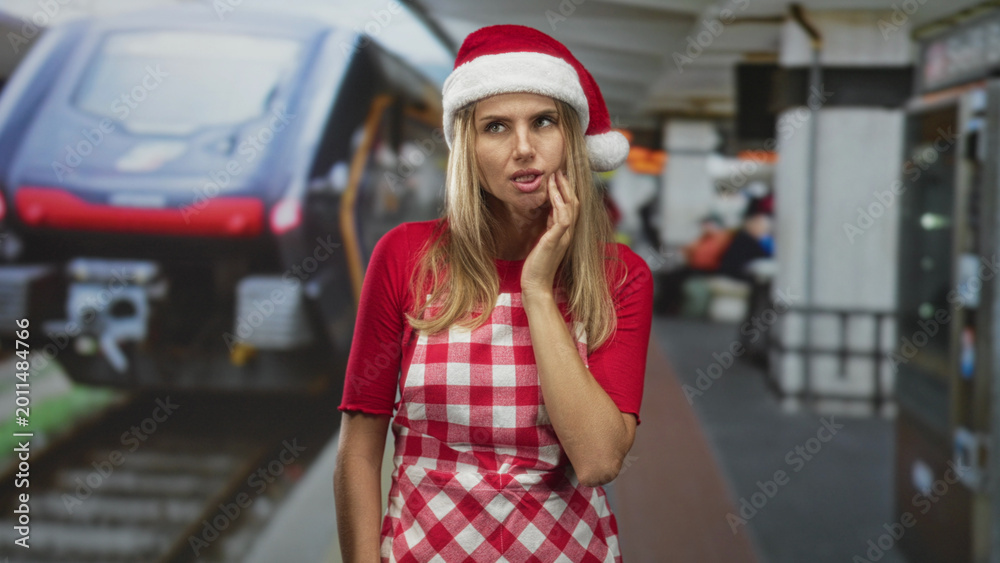 Naklejka premium Blonde woman wearing santa hat and checkered apron with hand on cheek for toothache at train platform inside a station building; holiday pain.