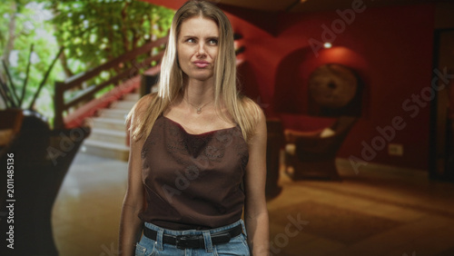 Schilderij op canvas Woman puckers lips beside staircase in building, wearing brown camisole and denim jeans, shoulders exposed and arms relaxed at sides; skepticism