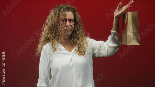 Fotografi Woman middle age holding a paper gift bag with heart in hand and raising the other hand in studio; disappointment contemplation