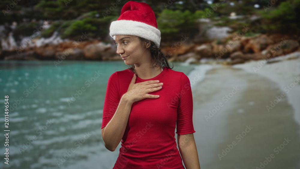 Naklejka premium Young woman in red shirt wearing santa hat places hand on chest on a tropical beach, eyes closed in a reflective pose; quiet gratitude.