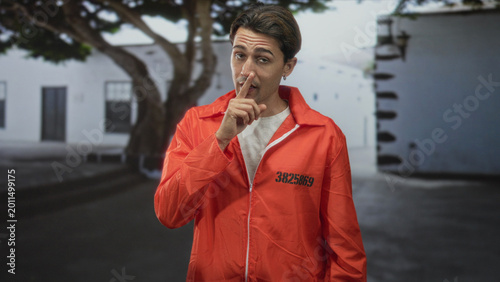 Canvas Print Man prisoner in orange jumpsuit, finger to lips for silence in prison building courtyard with inmate number visible; secrecy