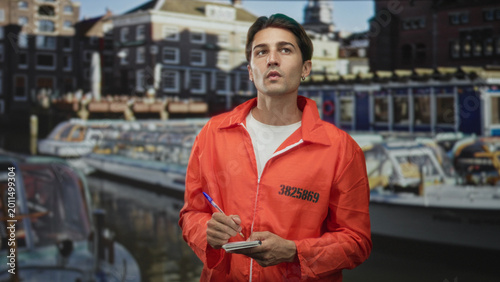 Foto Man in orange inmate jumpsuit writing with a pen on a notepad by canal building and boats; remorse reflection