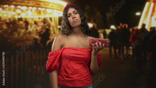Tableau sur toile Woman holding strawberries in a clear plastic punnet with bare shoulder exposed, hand raised in a presenting gesture at a street carnival at night; melancholy solitude