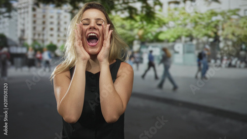 Fotografie Woman shouting with hands cupped to mouth on urban street with buildings and trees visible; joy celebration public expression