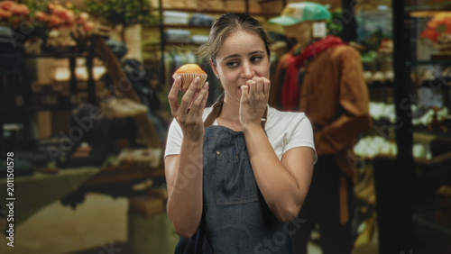 Bild auf Leinwand Young woman holding a cupcake and biting her fingernails in a retail building near mannequins and racks while wearing a denim apron; nervous hesitation