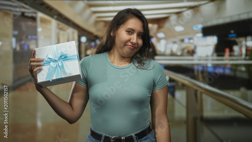 Fotografija Woman holding a gift box aloft with one hand and slight shrug by a mall railing inside a modern building; amused