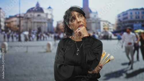 Bild auf Leinwand Woman holding makeup brushes with hand on chin in street, city square visible behind her; contemplation creativity