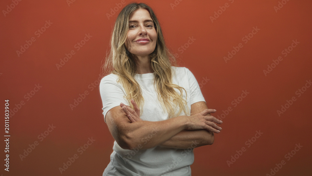 Obraz premium Woman smiling and posing, gesturing with a blurred hand and later folding crossed arms while wearing a white t shirt against an orange studio backdrop; confidence warmth.