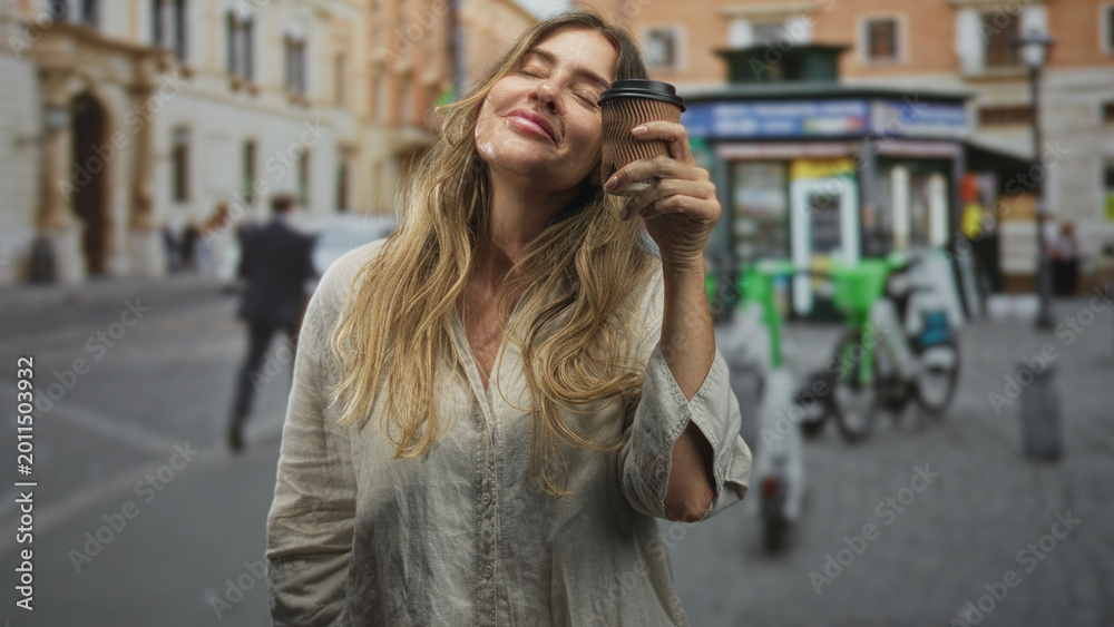 Fototapeta premium Woman holding coffee cup to face, eyes closed and smiling on a cobblestone street; contentment morning ritual.