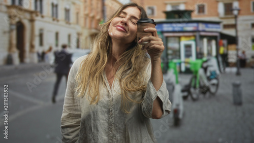 Tableau sur toile Woman holding coffee cup to face, eyes closed and smiling on a cobblestone street; contentment morning ritual