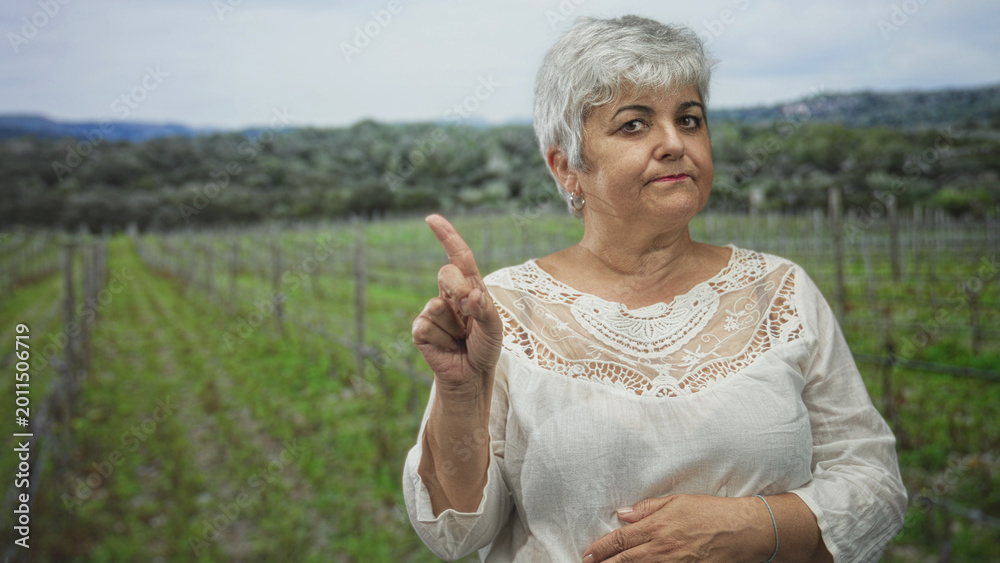 Obraz premium Woman, senior with grey hair, points finger and holds hand on stomach in a vineyard forest, wearing white lace blouse and hoop earring; firm admonition.